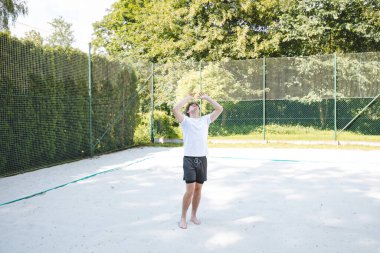 Genç bir adam güneşli bir günde kumlu bir sahada voleybol topunu havaya fırlatıyor. Günlük açık hava sahnesinde eğlence, yaz sporları ve doğal ortamlarda dinlenme vurgulanıyor..
