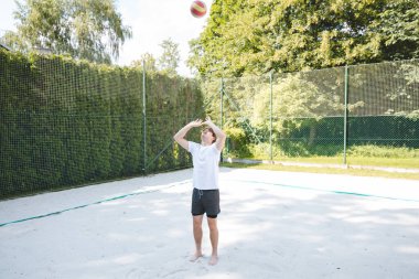 Genç bir adam güneşli bir günde kumlu bir sahada voleybol topunu havaya fırlatıyor. Günlük açık hava sahnesinde eğlence, yaz sporları ve doğal ortamlarda dinlenme vurgulanıyor..