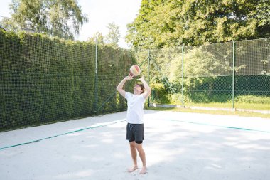 Genç bir adam güneşli bir günde kumlu bir sahada voleybol topunu havaya fırlatıyor. Günlük açık hava sahnesinde eğlence, yaz sporları ve doğal ortamlarda dinlenme vurgulanıyor..