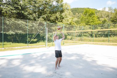 Genç bir adam güneşli bir günde kumlu bir sahada voleybol topunu havaya fırlatıyor. Günlük açık hava sahnesinde eğlence, yaz sporları ve doğal ortamlarda dinlenme vurgulanıyor..