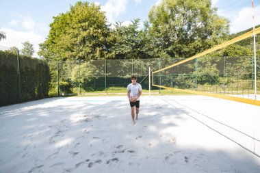 Genç adam, güneşli bir günde voleybol sahasında voleybol oynamaya hazırlanıyor. Yaz sporları, fitness ve açık hava eğlencesi sahneleri için ideal..