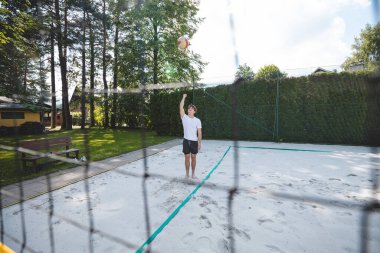 Genç bir adam voleybol topunu havaya fırlatıyor, bir kum sahası kazığına hazırlanıyor. Kalenin arkasından çekilen, dinamik bir açık hava sporu anı sergiliyor..
