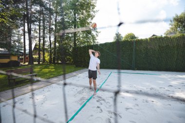 Genç bir adam voleybol topunu havaya fırlatıyor, bir kum sahası kazığına hazırlanıyor. Kalenin arkasından çekilen, dinamik bir açık hava sporu anı sergiliyor..