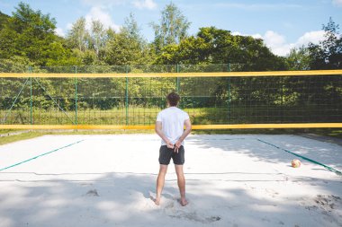 Voleybol oyuncusu voleybol sahasında duruyor, elleri arkada, bir sonraki oyuna hazırlanıyor. Açık hava voleybol maçının ortasında sakin bir an..