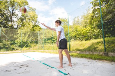 Voleybol oyuncusu kumda pas pratiği yapıyor, teknik ve kontrole odaklanıyor. Sahilde bir voleybol sahasında yakalanmış..