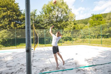 Voleybol oyuncusu bir açık hava maçı sırasında topu ağdan atmak için atlıyor. Eylem, plaj voleybolunun yoğunluğunu ve atletizmini yakalıyor..