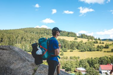Mavi sırt çantalı bir yürüyüşçü kayanın üzerinde duruyor, yemyeşil ormanlara tepeden bakan ve Beskydy Dağları 'ndaki parlak mavi gökyüzünün altında dolambaçlı bir yol, manzaranın tadını çıkarıyor..