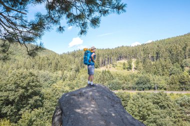 Hiker with a blue backpack stands atop a rock, overlooking lush green forests and a winding path under a bright blue sky in the Beskydy Mountains, enjoying the view. clipart