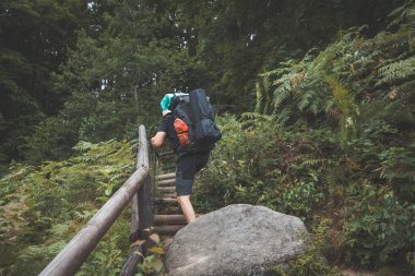 Mavi sırt çantalı bir gezgin, yosun kaplı kayalar ve yoğun yeşilliklerle çevrili metal merdivenlerden Beskydy Dağları 'ndaki engebeli bir orman yolunda dikkatle tırmanır..
