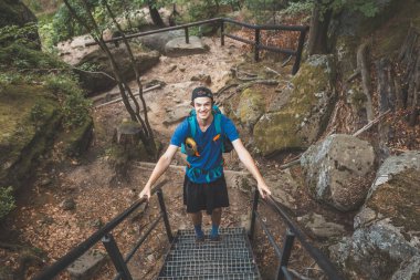 Mavi sırt çantalı bir gezgin, yosun kaplı kayalar ve yoğun yeşilliklerle çevrili metal merdivenlerden Beskydy Dağları 'ndaki engebeli bir orman yolunda dikkatle tırmanır..