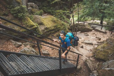 Hiker with a blue backpack carefully ascends metal stairs surrounded by moss-covered rocks and dense greenery, navigating a rugged forest trail in the Beskydy Mountains. clipart