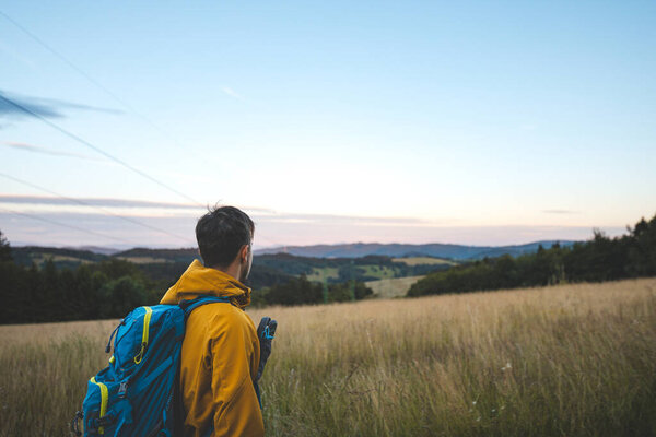 Hiker in a yellow jacket with a blue backpack overlooking a peaceful landscape at sunset in the Beskydy Mountains, Czech Republic. A quiet moment of reflection and nature.