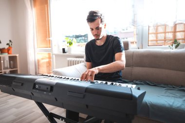 Adam, modern dekor ve sıcak doğal ışıkla çevrili, güneşli bir oturma odasında dijital bir klavye çalmaya odaklanmış. Huzurlu ve ilham verici bir atmosfer yaratıyor..