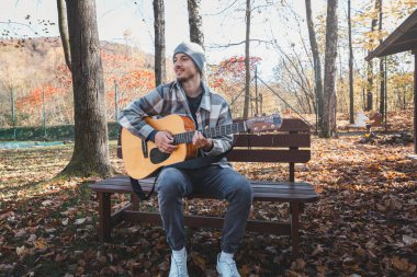 Young man sitting on a bench in a scenic autumn park, playing an acoustic guitar, surrounded by colorful trees and fallen leaves. Capturing calmness, creativity, and nature. clipart