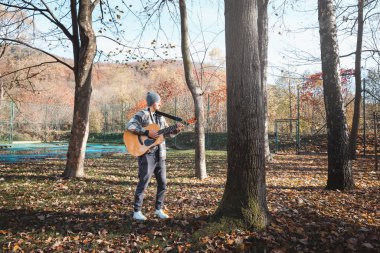 Ekose gömlek giyen ve bereli bir adam sakin bir sonbahar parkında durup akustik gitar tıngırdatıyor. Renkli yapraklar ve uzun ağaçlarla çevrili açık havada müzik için yaratıcılığını ve tutkusunu ifade eder..