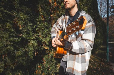 Ekose gömlekli odaklanmış genç bir adam ve bereli bir akustik gitar çalıyor yemyeşil çalıların arasında. Müzik tutkusu ve doğa ile bağlantı kurma sahnesi..