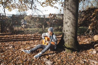 young man in a plaid shirt and beanie sits under a large tree, playing an acoustic guitar in a sunny autumn setting. The ground is covered with dry leaves, enhancing the cozy and peaceful atmosphere. clipart