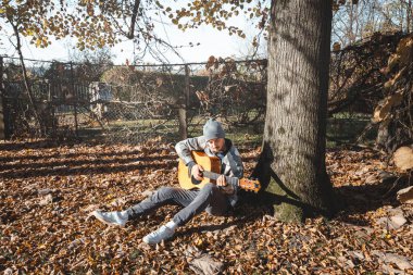Ekose gömlekli ve bereli genç adam güneşli bir sonbahar ortamında büyük bir ağacın altında akustik gitar çalıyor. Zemin kuru yapraklarla kaplı. Sıcak ve huzurlu atmosferi arttırıyor..
