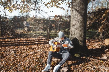 Ekose gömlekli ve bereli genç adam güneşli bir sonbahar ortamında büyük bir ağacın altında akustik gitar çalıyor. Zemin kuru yapraklarla kaplı. Sıcak ve huzurlu atmosferi arttırıyor..