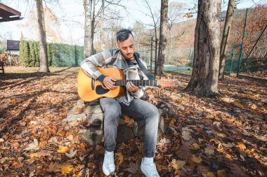 Ekose gömlekli odaklanmış gitarist, altın sonbahar manzarasının ortasında taş bir bankta otururken akustik gitar çalar. Güneş ışığı, sıcak ve huzurlu ortamı güçlendirir..