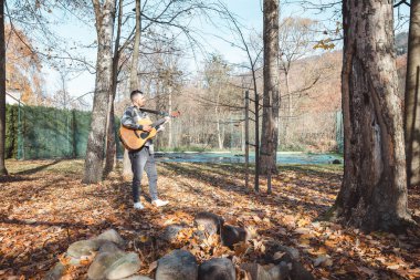 Ekose gömlekli gitarist, sonbahar parkındaki bir taş yığınının üzerinde güvenle duruyor, akustik gitarını tıngırdatıyor. Altın yapraklar ve uzun ağaçlarla çevrili, yaratıcılığı ve sükuneti somutlaştırıyor..