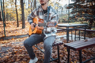 Genç bir adam ağaca yaslanmış, güneşli bir sonbahar parkında akustik gitar çalıyor, yapraklarla ve doğal renklerle çevrili, dingin bir atmosfer yaratıyor.