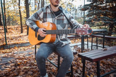 Genç bir adam ağaca yaslanmış, güneşli bir sonbahar parkında akustik gitar çalıyor, yapraklarla ve doğal renklerle çevrili, dingin bir atmosfer yaratıyor.