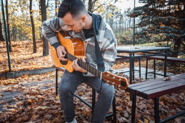 Genç bir adam ağaca yaslanmış, güneşli bir sonbahar parkında akustik gitar çalıyor, yapraklarla ve doğal renklerle çevrili, dingin bir atmosfer yaratıyor.