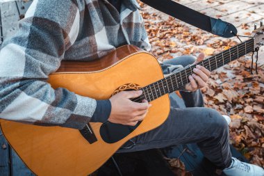 Genç bir adam ağaca yaslanmış, güneşli bir sonbahar parkında akustik gitar çalıyor, yapraklarla ve doğal renklerle çevrili, dingin bir atmosfer yaratıyor.