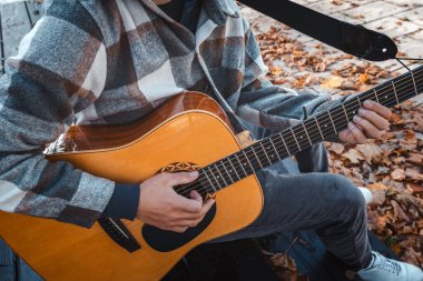 Genç bir adam ağaca yaslanmış, güneşli bir sonbahar parkında akustik gitar çalıyor, yapraklarla ve doğal renklerle çevrili, dingin bir atmosfer yaratıyor.
