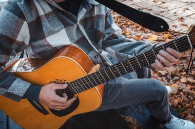 Genç bir adam ağaca yaslanmış, güneşli bir sonbahar parkında akustik gitar çalıyor, yapraklarla ve doğal renklerle çevrili, dingin bir atmosfer yaratıyor.