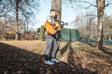 Young man leaning against a tree, playing an acoustic guitar in a sunlit autumn park, surrounded by fallen leaves and natures vibrant colors, creating a serene atmosphere clipart