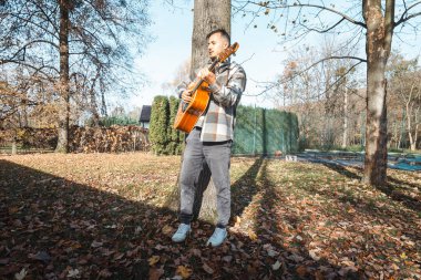 Young man leaning against a tree, playing an acoustic guitar in a sunlit autumn park, surrounded by fallen leaves and natures vibrant colors, creating a serene atmosphere clipart