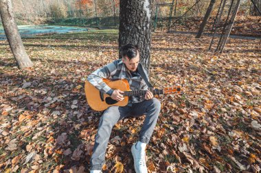 Ekose gömlekli bir adam, sonbahar yaprakları üzerinde sakin bir parkta otururken akustik gitarını akort eder. Canlı sonbahar renkleri ve doğal ortam yaratıcı havayı arttırır..