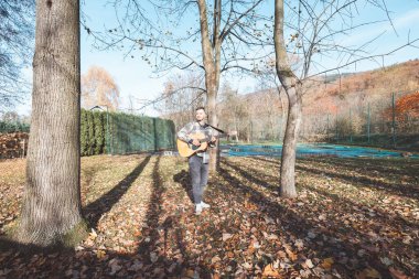 Müzisyen, sonbaharda düşen yapraklar ve doğal güzelliklerle çevrili güneşli bir parkta akustik gitar çalar. Canlı ortam müzik performansını tamamlıyor..