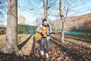 Müzisyen, sonbaharda düşen yapraklar ve doğal güzelliklerle çevrili güneşli bir parkta akustik gitar çalar. Canlı ortam müzik performansını tamamlıyor..