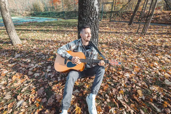 Ekose gömlekli bir adam, sonbahar yaprakları üzerinde sakin bir parkta otururken akustik gitarını akort eder. Canlı sonbahar renkleri ve doğal ortam yaratıcı havayı arttırır..