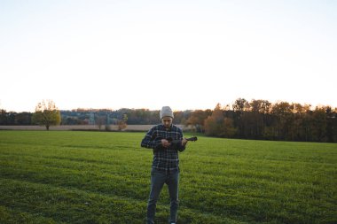 Ekose gömlekli bir müzisyen ve bereli gün batımında yeşil bir tarlada ukulele çalıyor. Sakin kırsal sahne, müzik, doğa ve sükuneti güzel bir şekilde birleştirir..