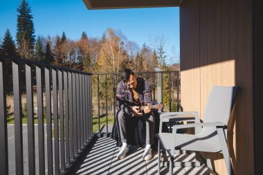 Young musician sits on a sunny balcony, immersed in playing the ukulele. Surrounded by autumn trees, he enjoys the peaceful moment outdoors, lost in his thoughts and music clipart