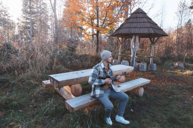 Müzisyen, günışığıyla aydınlatılmış bir ormandaki ahşap bir bankta oturup ukulele çalar. Sonbahar yaprakları ve ağaçların arasından akan ılık güneş ışığı ile çevrili..