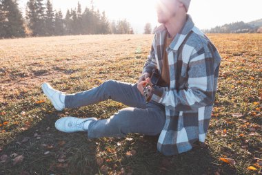 Genç adam sıcak sonbahar günışığında çimenli bir tarlada oturuyor, ukulele çalarken gülümsüyor. Altın tonlar ve uzak ağaçlarla çevrili bu an neşeyi, yaratıcılığı ve derin bir bağlantıyı yansıtıyor..