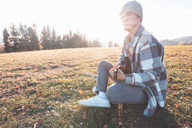 Genç adam sıcak sonbahar günışığında çimenli bir tarlada oturuyor, ukulele çalarken gülümsüyor. Altın tonlar ve uzak ağaçlarla çevrili bu an neşeyi, yaratıcılığı ve derin bir bağlantıyı yansıtıyor..