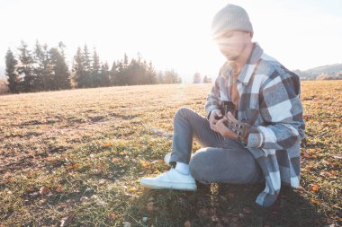Genç adam sıcak sonbahar günışığında çimenli bir tarlada oturuyor, ukulele çalarken gülümsüyor. Altın tonlar ve uzak ağaçlarla çevrili bu an neşeyi, yaratıcılığı ve derin bir bağlantıyı yansıtıyor..