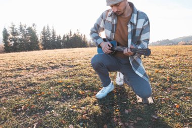 Genç adam sıcak sonbahar günışığında çimenli bir tarlada oturuyor, ukulele çalarken gülümsüyor. Altın tonlar ve uzak ağaçlarla çevrili bu an neşeyi, yaratıcılığı ve derin bir bağlantıyı yansıtıyor..