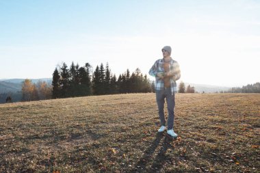 Musician stands in an open field during a sunny autumn day, holding a ukulele. Surrounded by hills and trees, the serene scene reflects freedom, creativity, and a connection to nature. clipart