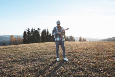 Musician stands in an open field during a sunny autumn day, holding a ukulele. Surrounded by hills and trees, the serene scene reflects freedom, creativity, and a connection to nature. clipart