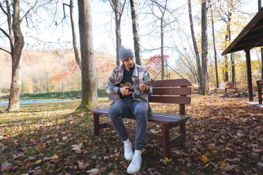 Tutku, sonbahar yaprakları arasında parktaki ahşap bir bankta oturup ukulele çalıyor. Etrafı ağaçlarla çevrili ve yumuşak güneş ışığıyla yıkanmış, yaratıcılıkla dolu huzurlu bir anın tadını çıkarıyor..