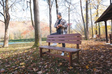 Tutku, sonbahar yaprakları arasında parktaki ahşap bir bankta oturup ukulele çalıyor. Etrafı ağaçlarla çevrili ve yumuşak güneş ışığıyla yıkanmış, yaratıcılıkla dolu huzurlu bir anın tadını çıkarıyor..