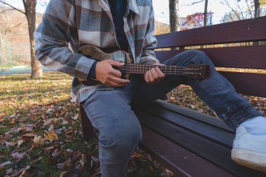 Tutku, sonbahar yaprakları arasında parktaki ahşap bir bankta oturup ukulele çalıyor. Etrafı ağaçlarla çevrili ve yumuşak güneş ışığıyla yıkanmış, yaratıcılıkla dolu huzurlu bir anın tadını çıkarıyor..