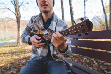 Tutku, sonbahar yaprakları arasında parktaki ahşap bir bankta oturup ukulele çalıyor. Etrafı ağaçlarla çevrili ve yumuşak güneş ışığıyla yıkanmış, yaratıcılıkla dolu huzurlu bir anın tadını çıkarıyor..